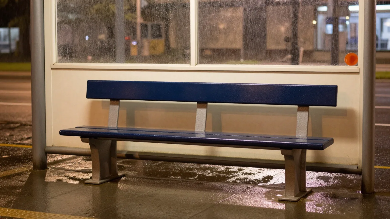 Eine leere Bank an einer Straßenbahnhaltestelle bei Regen in der Nacht, nasses Pflaster spiegelt die Straßenlaternen