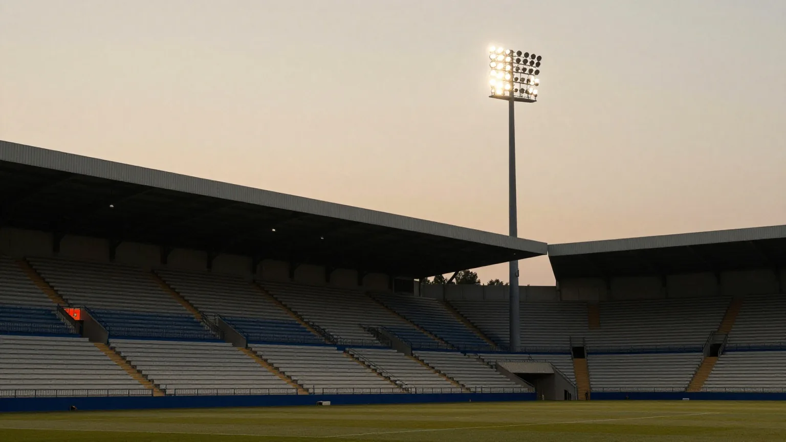 Hohe Flutlichtmasten leuchten über einem leeren Fußballplatz in der Abenddämmerung, Reihen leerer Sitze im schwachen Abendlicht