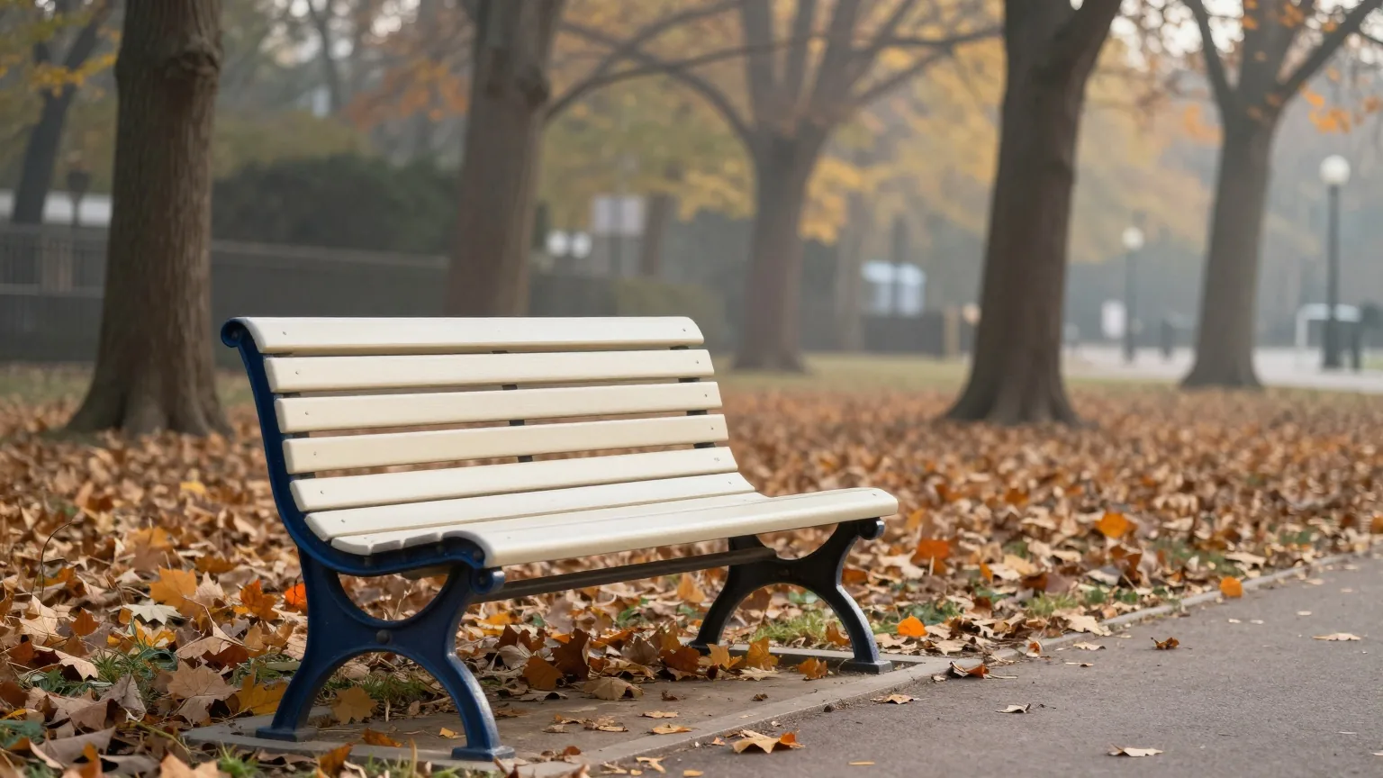 Leere Holzbank an einem von Bäumen gesäumten Parkweg, Herbstlaub auf dem Boden, frühes Morgenlicht