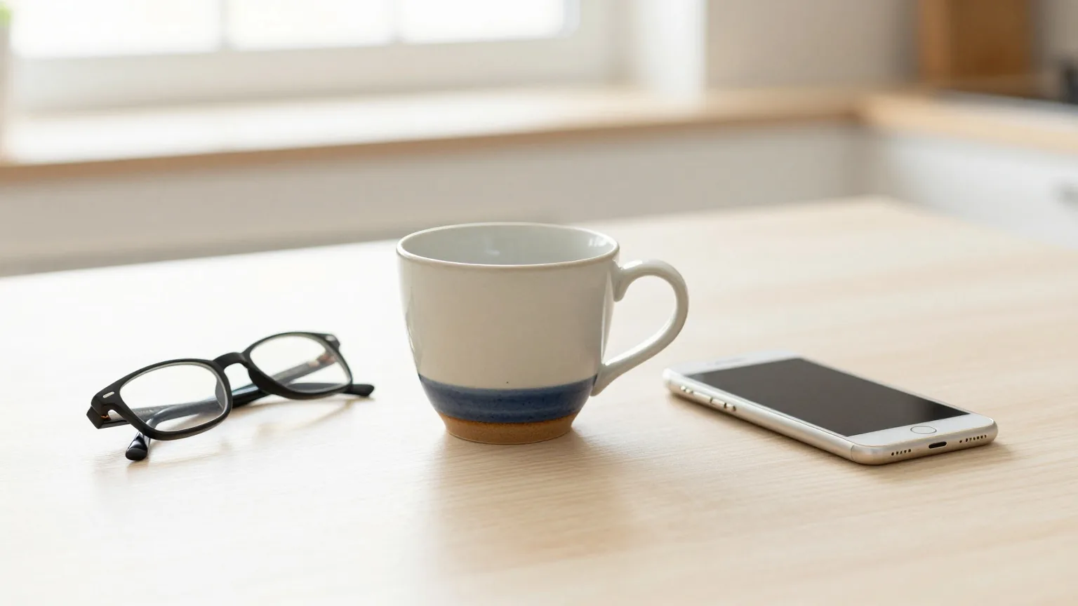 Heller Küchentisch am frühen Morgen mit leerer Keramiktasse, einer Lesebrille und einem Smartphone mit dem Bildschirm nach unten