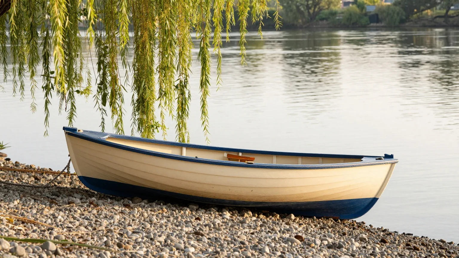 Hölzernes Ruderboot am Kieselufer eines ruhigen Flusses, tief hängende Weidenzweige über dem Wasser im warmen Licht des späten Nachmittags