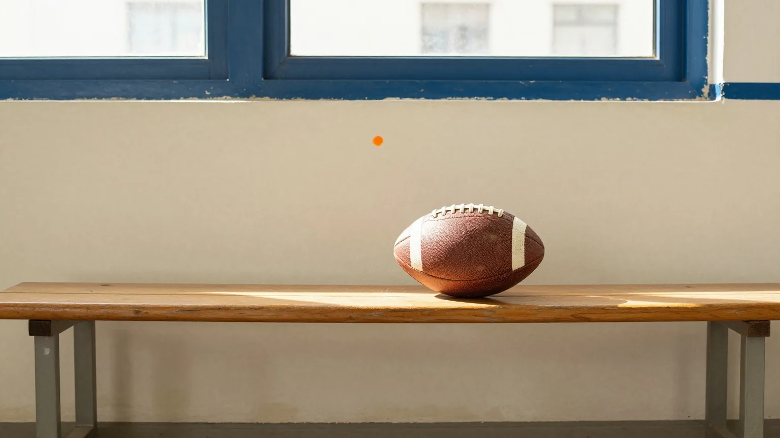 Ein abgewetzter Lederfußball unter einer Holzbank an der Wand einer leeren Schulsporthalle, Staubkörner im schrägen Nachmittagslicht