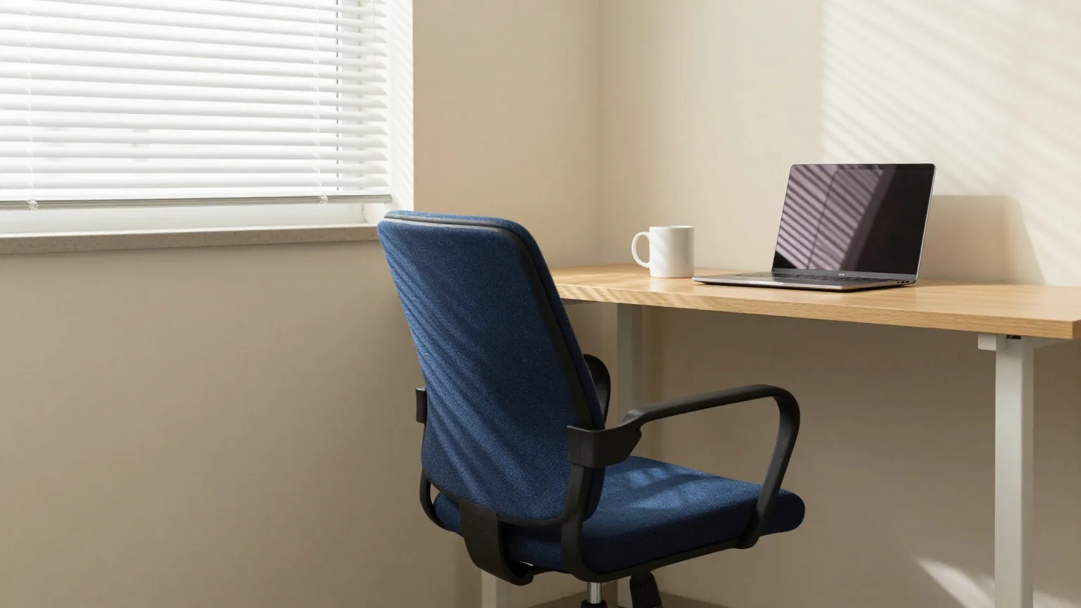 Ruhige Ecke eines kleinen Gemeinschaftsbüros mit einem unter den Holzschreibtisch geschobenen Drehstuhl, daneben eine weiße Tasse und ein zugeklappter Laptop, Nachmittagslicht durch eine Jalousie