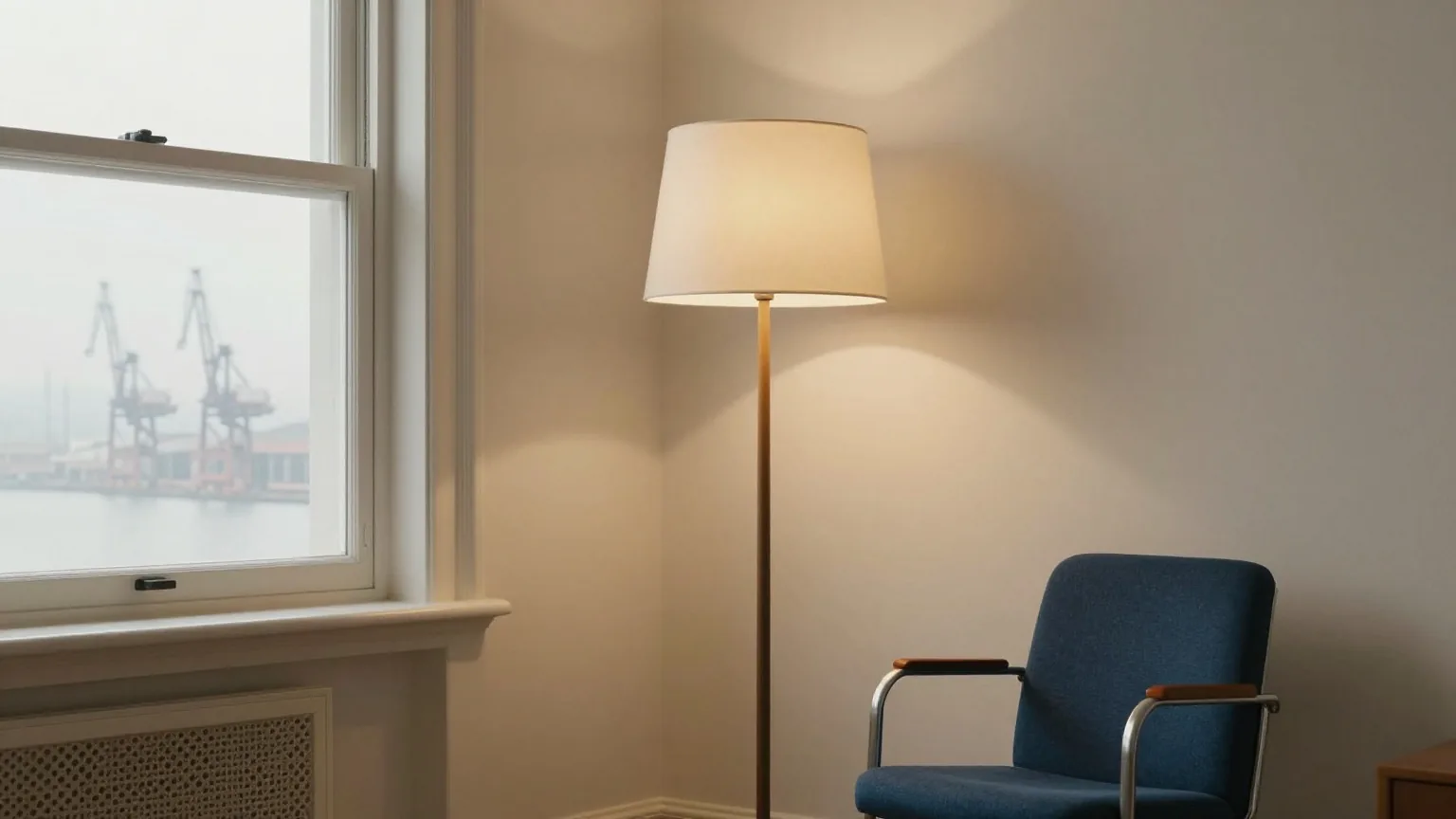 Ausgeschaltete Stehlampe neben einem leeren Lesesessel in einem Altbau, draußen Hafenkräne im Morgendunst
