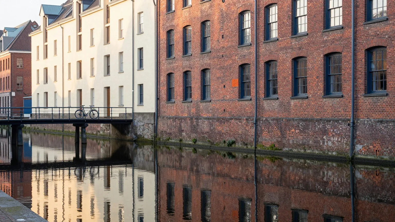 Rote Backsteinspeicher spiegeln sich im ruhigen Kanalwasser im Morgengrauen, ein Fahrrad lehnt am Brückengeländer