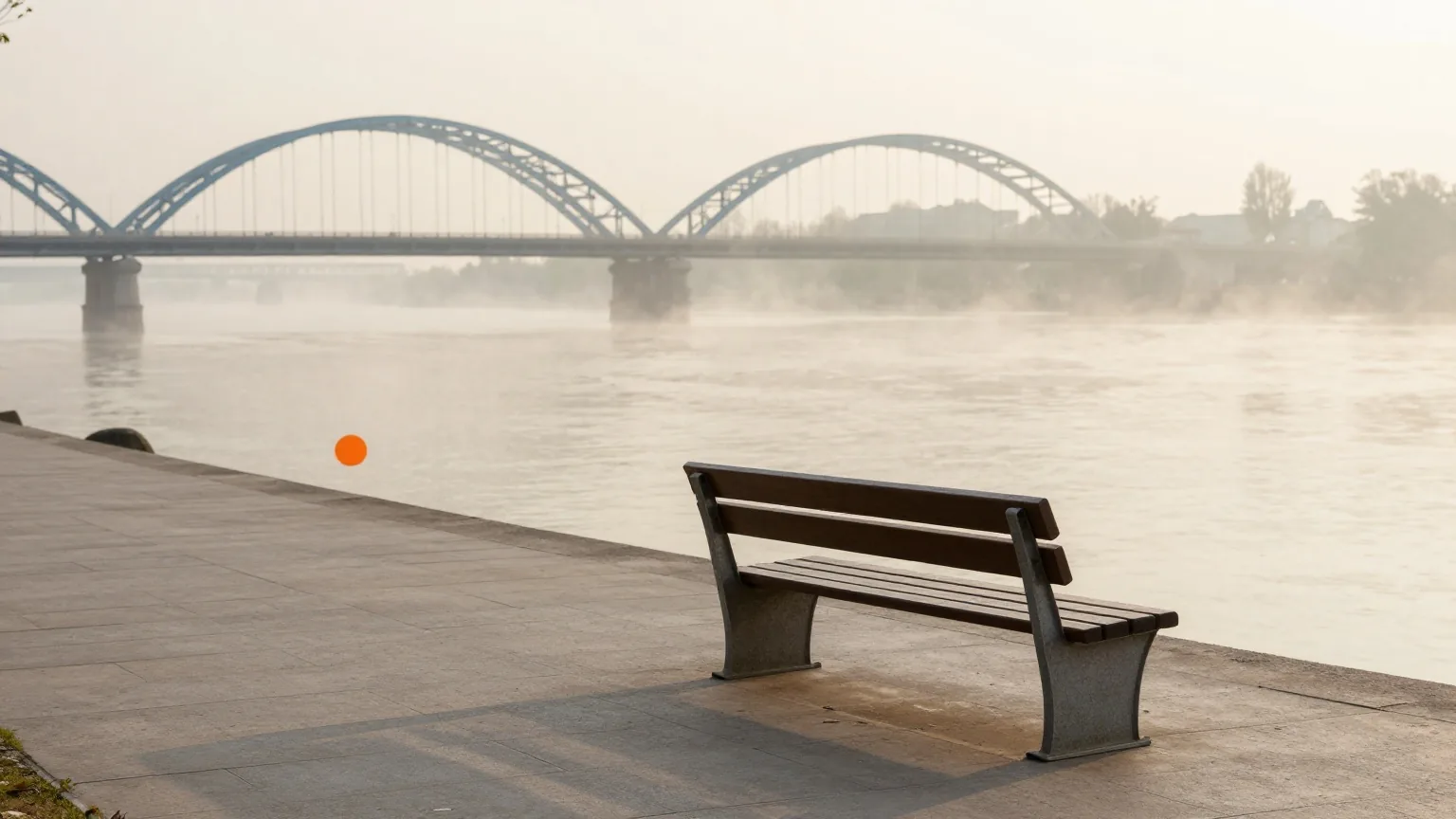 Leere Uferpromenade mit einer Holzbank im Morgengrauen, Nebel über dem breiten Fluss und Bögen einer Stahlbrücke in der Ferne