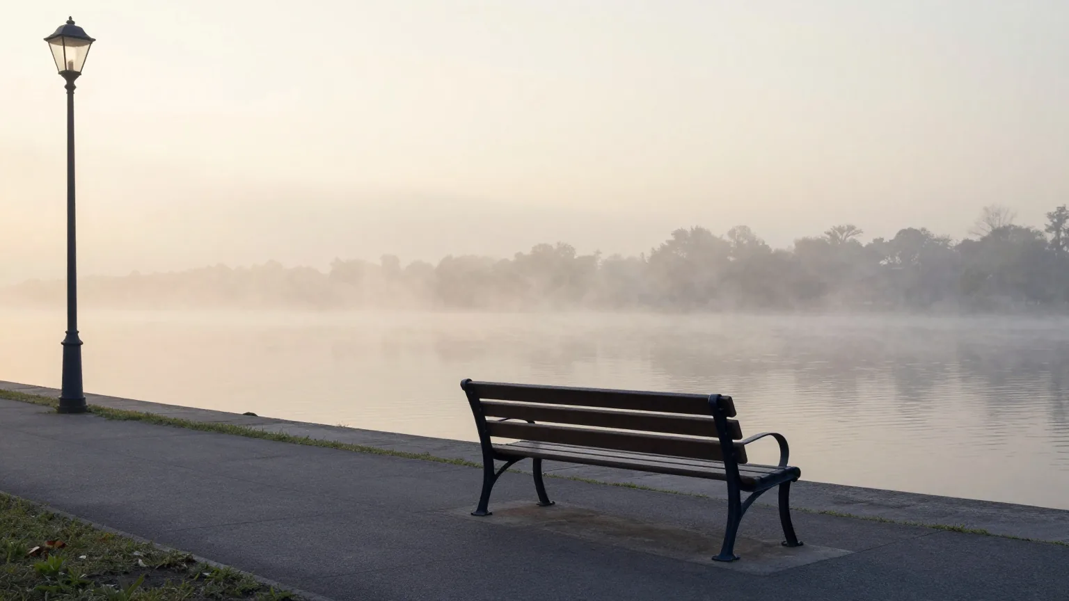 Leere Bank an einer Flusspromenade im Morgengrauen, Nebel über dem Wasser, einige Laternen brennen noch, Weitwinkel aus niedriger Position