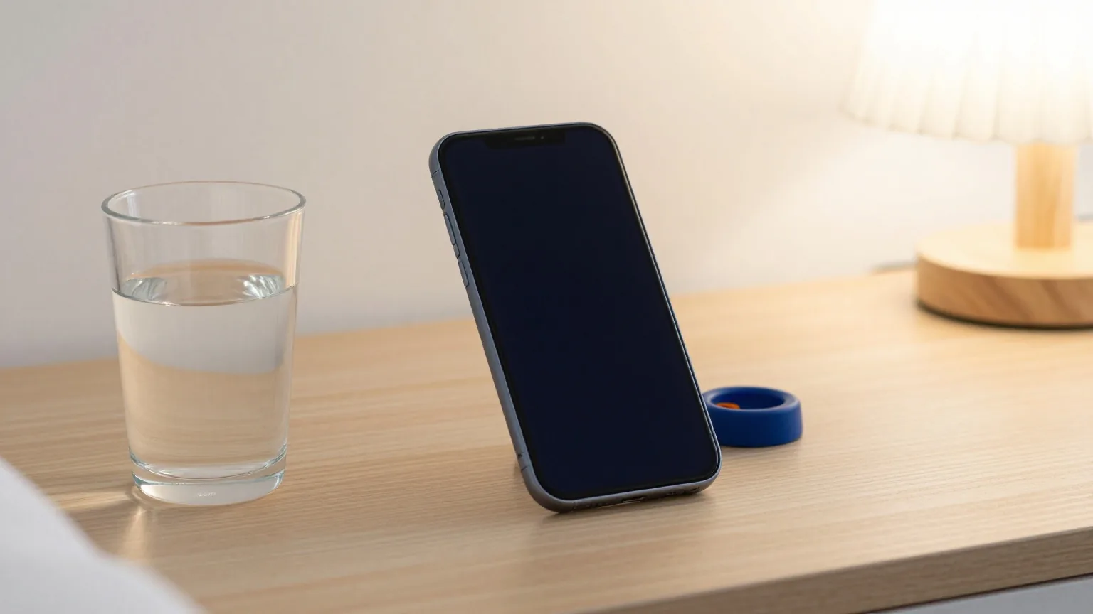Smartphone mit ausgeschaltetem Display auf einem hölzernen Nachttisch neben einem Wasserglas und einer ausgeschalteten Lampe, weiches Morgenlicht durch einen Vorhang