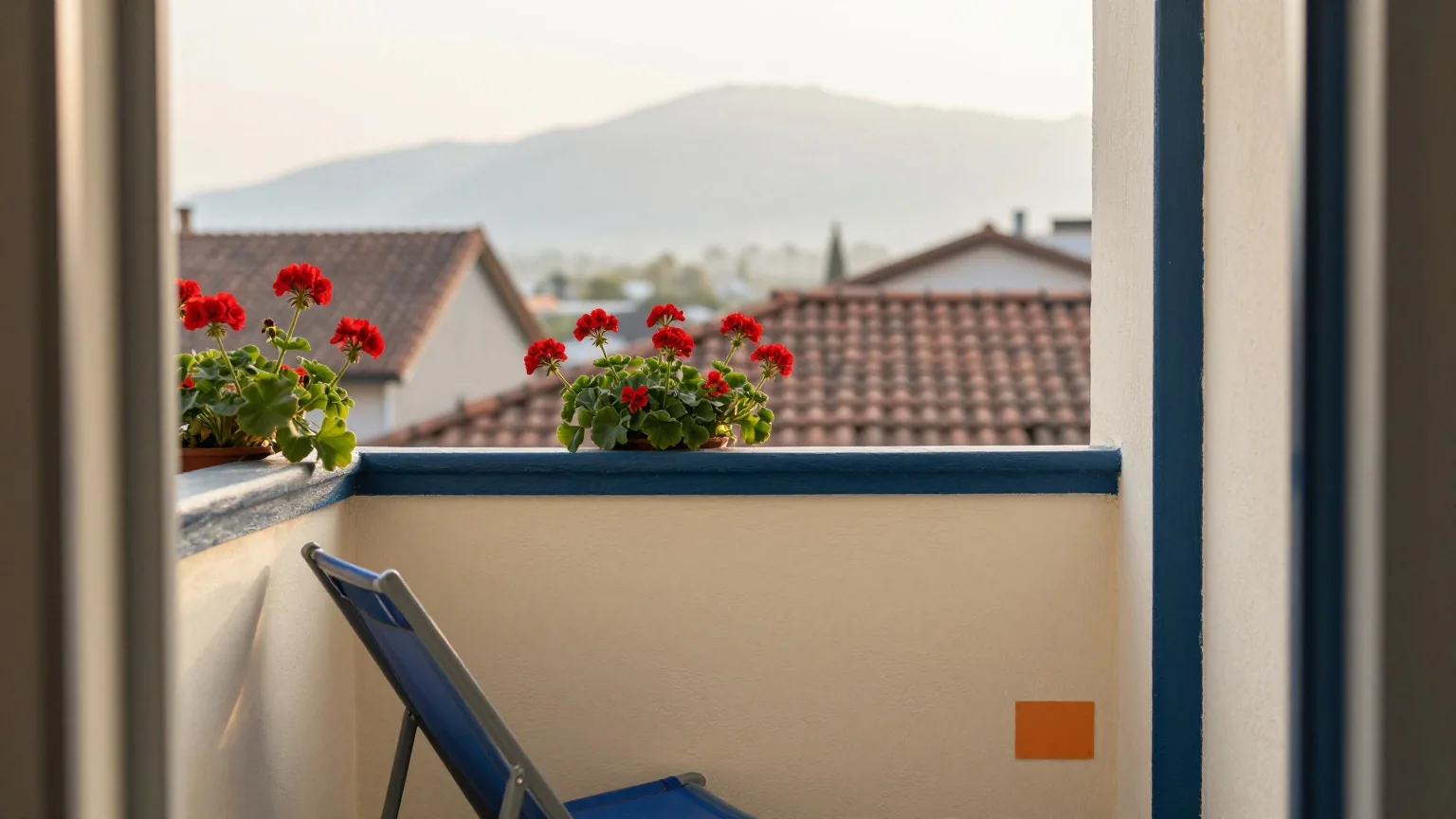 Stiller Stadtbalkon mit roten Geranien und zusammengeklapptem Liegestuhl, in der Ferne Berge im Morgendunst über Ziegeldächern