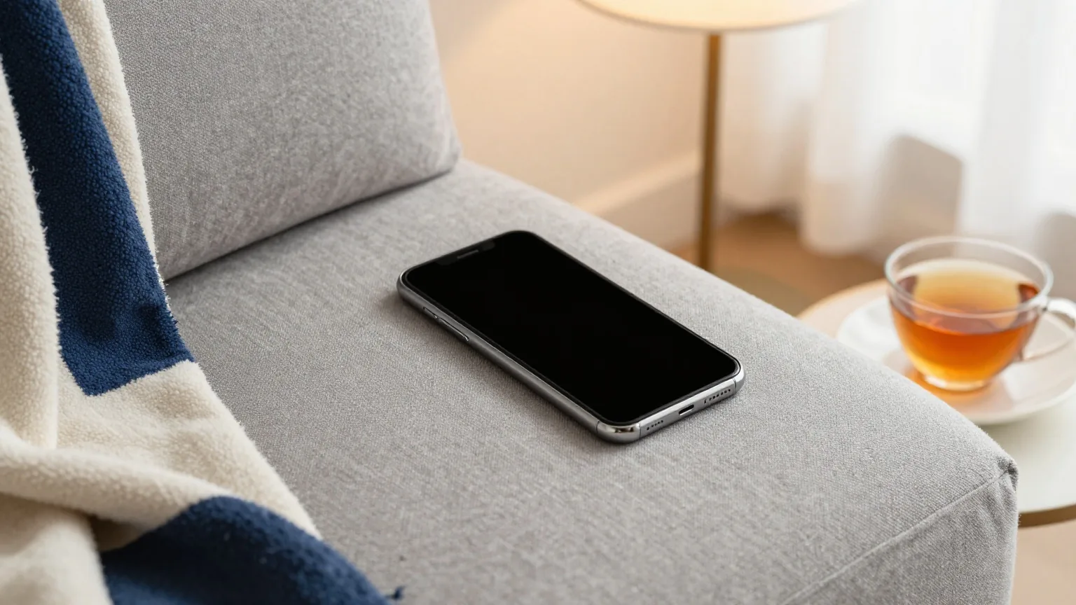 Smartphone mit ausgeschaltetem Display auf einem grauen Leinenkissen neben einer gefalteten Decke und einer Tasse Tee, weiches Abendlicht einer Stehlampe