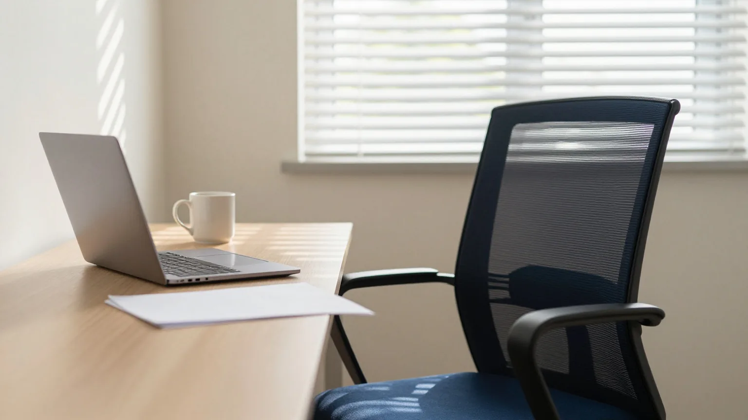 Leerer Bürostuhl an einem hellen Holzschreibtisch mit zugeklapptem Laptop, gefaltetem Blatt Papier und Tasse im Streifenlicht der Jalousie