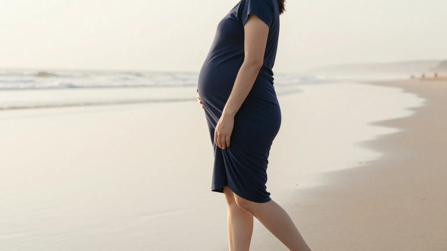 Schwangere Frau von hinten am Strand bei Ebbe, eine Hand auf dem Bauch, im blassen Morgenlicht